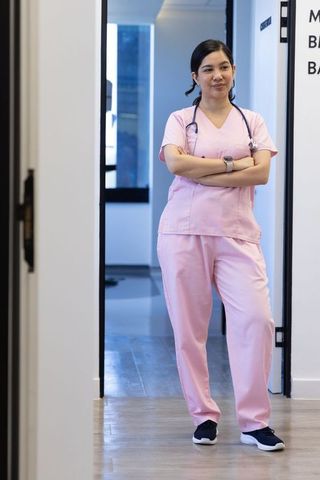 Confident nurse standing in doorway wearing pink scrubs and stethoscope, crossing arms