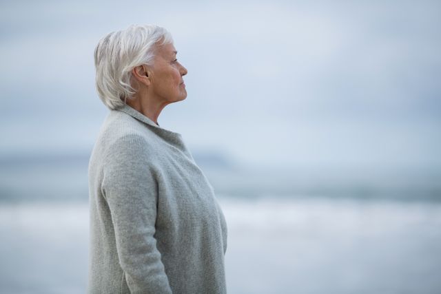 Senior woman standing on a beach, smiling and enjoying the peaceful ocean view. Ideal for use in advertisements or articles related to retirement, relaxation, mental health, and outdoor activities for seniors.