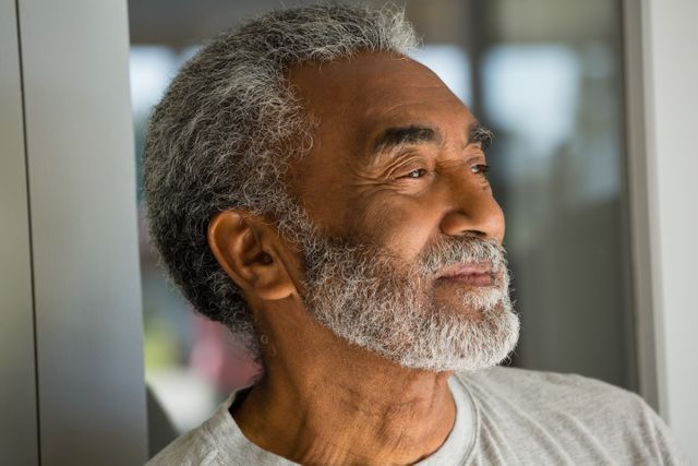 This image shows a happy senior man standing at the entrance of his house, smiling and looking content. The natural light highlights his grey hair and beard, giving a sense of warmth and relaxation. This image can be used in advertisements for retirement communities, senior living, healthcare services, or lifestyle blogs focusing on aging gracefully.