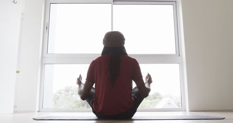 African american man meditating in minimalist home, sitting cross-legged facing window
