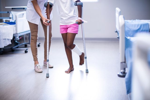 Female doctor assisting girl to walk with crutches in ward of hospital