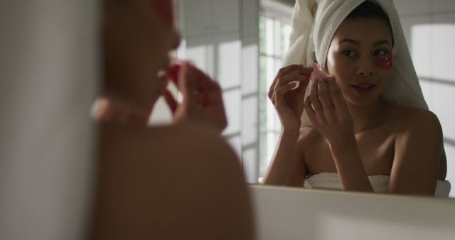Vertical image of smiling biracial woman with towel on hair applying face mask in bathroom. Health and beauty, leisure time, domestic life and lifestyle concept.