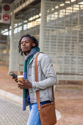African american commuter waiting at bus stop holding coffee and smartphone urban commute