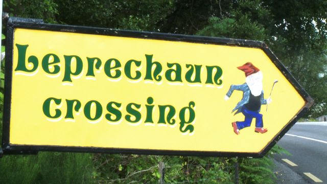 Sign depicting 'Leprechaun Crossing' set outdoors, surrounded by green trees and foliage. Ideal for illustrating themes of Irish folklore, humor in travel, or fantasy-themed content. Useful for articles on cultural symbols, local tourism, or roadside curiosities worldwide.