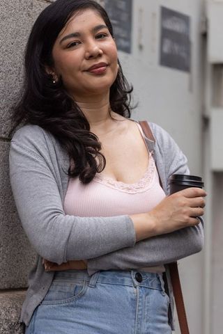 Urban woman leaning on stone wall holding takeaway coffee cup wearing cardigan and jeans