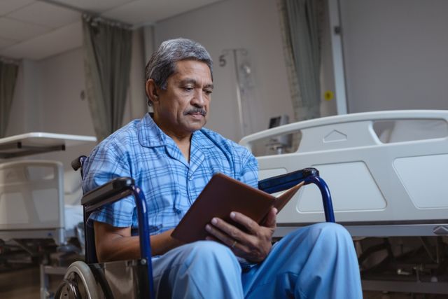 Front view of disabled mature male patient sitting on wheelchair and reading a book in medical ward at hospital