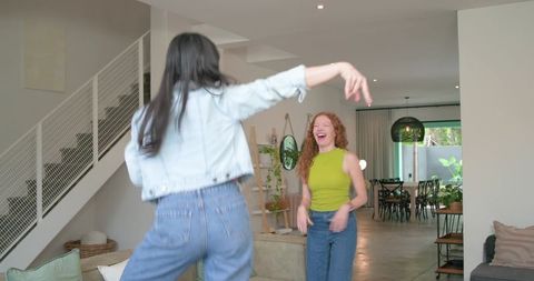 Two women jumping and laughing in modern living room wearing denim jacket and lime top