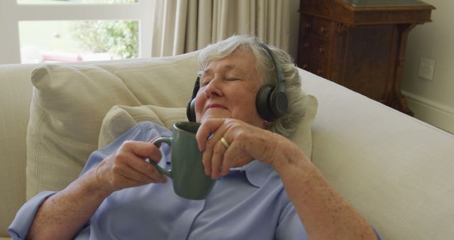 An elderly woman is reclining on a light colored couch, wearing headphones, and holding a coffee mug. She looks relaxed and possibly listening to music or a podcast. This image can be used to represent relaxation, leisure, retirement lifestyle, or senior living. Suitable for advertisements or articles related to elderly well-being, relaxation tips, or safe and comfortable home environments.