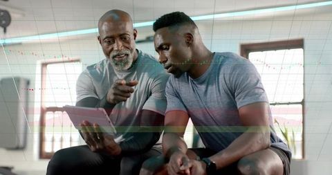 Personal trainer pointing at tablet reviewing workout plan with client on gym bench