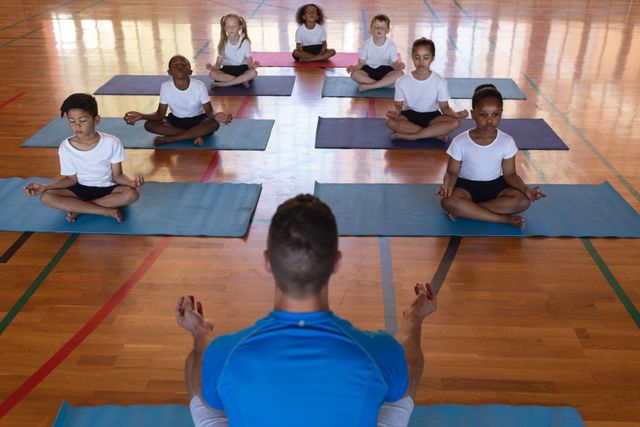 Yoga instructor leading a diverse group of children in meditation on yoga mats in a school gym. Ideal for use in educational materials, fitness and wellness promotions, and advertisements for children's health programs.