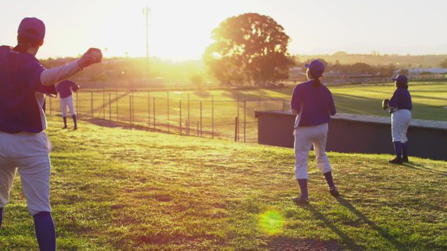 Group of female baseball players practicing by catching and throwing a ball during sunset. Field shows well-maintained grass and distant trees, giving an energetic atmosphere. Perfect for illustrating diversity in sports, women empowerment in athletics, or teamwork dynamics. Suitable for sport-related promotional material or community events related to baseball engagement.