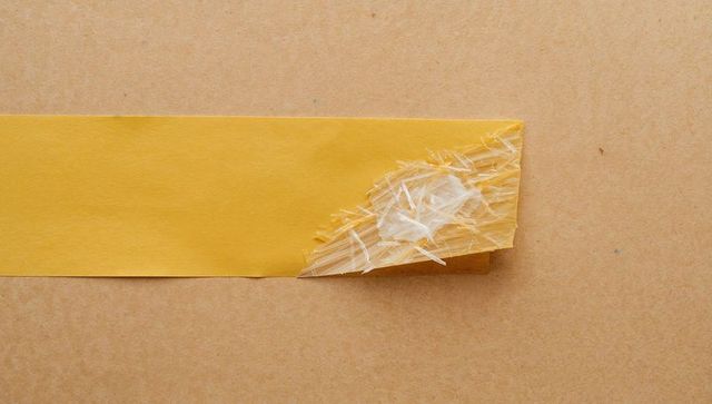 Peeling yellow masking tape revealing torn adhesive fibers on kraft cardboard strip detail