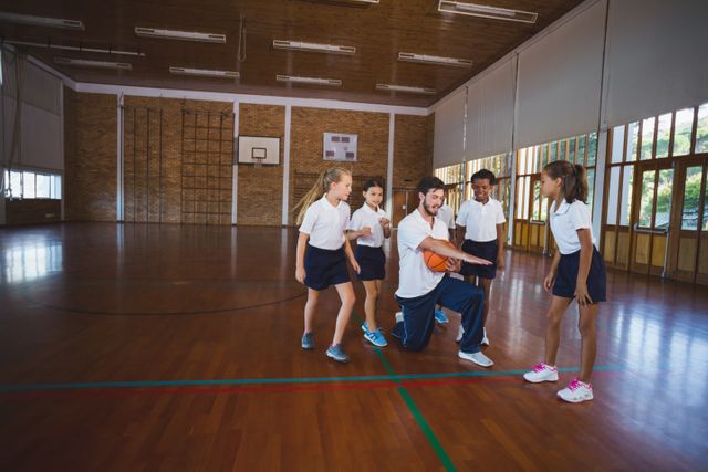 Sports teacher teaching school kids to play basketball in court at school gym