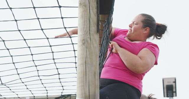 Strong and determined woman in pink sportswear climbing a net on an obstacle course at an outdoor event. Ideal for use in articles and promotions relating to fitness, outdoor activities, events, personal challenges, and women's empowerment.