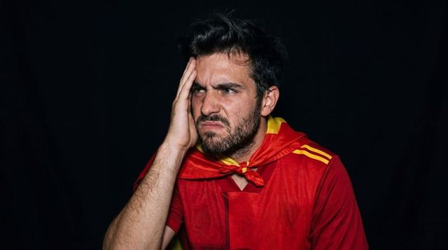 Frustrated sports fan holding head and grimacing wearing red cape with yellow trim