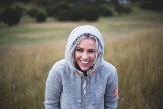 Smiling woman wearing gray hoodie in grassy field, capturing joyful outdoor moment. Perfect for campaigns about outdoor lifestyle, winter clothing, or portraying happiness and well-being. Suitable for blogs, social media, advertisements focusing on casual fashion or a healthy lifestyle.