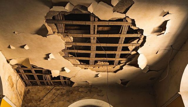 Peeling plaster ceiling revealing wooden lath and dangling wires in dilapidated hall