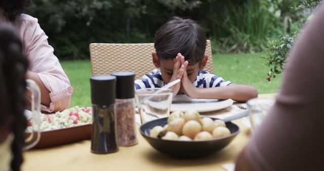 Biracial boy praying at table with meal in garden. Lifestyle, childhood, family, togetherness, food and domestic life, unaltered.