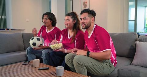 Friends watching soccer match on couch wearing red jerseys cheering with popcorn