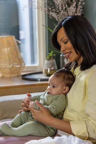 Asian mother cradling baby on sofa in sunlit living room, tender gentle bonding moment