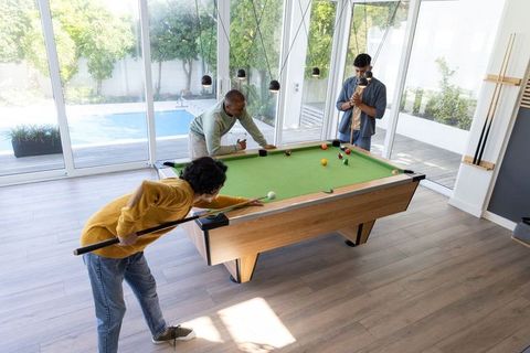 Multiracial friends playing pool in sunlit modern game room with glass doors and pool deck