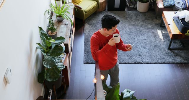 A man is sipping a cup of coffee while using his smartphone in a stylish, modern apartment. Various houseplants enhance the cozy ambiance. Great for content involving technology in everyday life, morning routines, home decor, relaxation, and lifestyle.