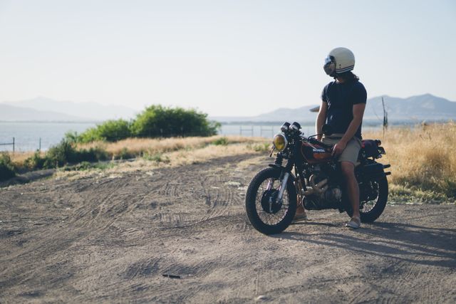 Man riding a vintage motorcycle on a dirt road near a lake with mountains in the background. Wearing a helmet and outdoor gear, expressing adventure and freedom. Ideal for banners, tourism ads, adventure magazines, blogs about motorcycle riding, travel experiences, and nature exploration.