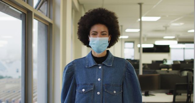 Young African woman in denim jacket wearing a mask standing in a modern, bright office. Ideal for use in content about COVID-19, workplace safety, modern office environments, and health protocols during the pandemic.