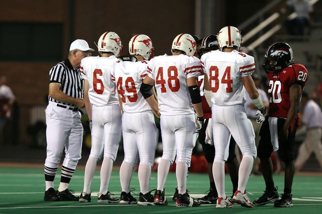 Several football players from two teams are standing in midfield facing the referee. The players in white uniforms are gathered together while the referee gives instructions. This can be used to illustrate football games, sportsmanship, team sports, and pre-game rituals.