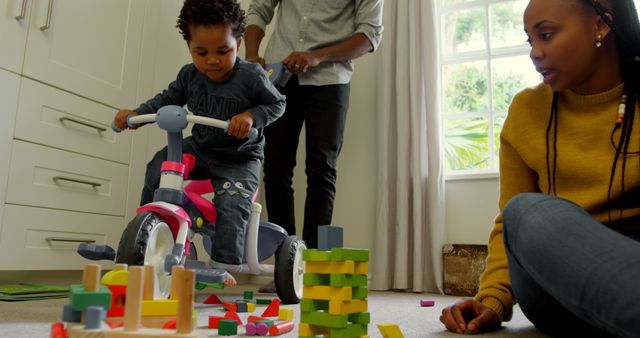 Parents engaging with toddler riding tricycle and stacking blocks in cozy living room. Ideal for family-oriented publications, parenting blogs, advertisements showcasing happy family life, and educational materials on childhood development and family bonding.