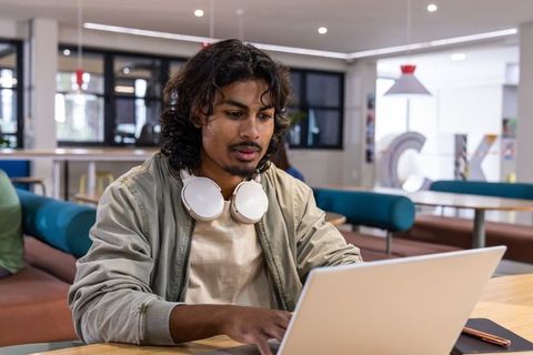 Young developer working on laptop in modern campus lounge with headphones around neck