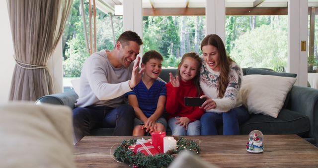 Family enjoying a moment together on the couch, waving at a smartphone during a video call. Setting includes Christmas decorations with candles and a wreath on the table, adding a festive touch. Used for promoting family bonding during holidays, festive greetings, and technology connecting loved ones.