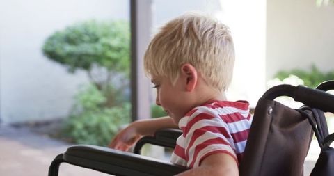 Young boy sitting in wheelchair on sunlit patio looking toward garden, candid mobility portrait