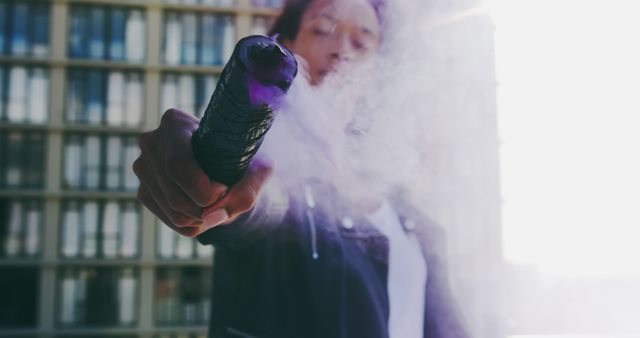 Woman holding a smoke grenade that is emitting purple smoke in an outdoor urban setting. Apartment buildings in the background against the bright sun create a vivid contrast. Image ideal for illustrating urban culture, street photography, event promotion materials, magazines focused on youth culture and modern urban activities.