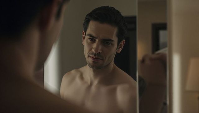 Shirtless man gazing into mirror in moody hotel room, contemplative intimate portrait