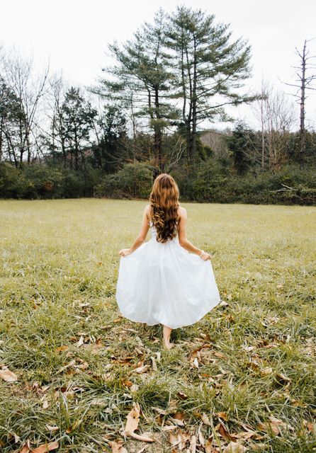 A young girl with long hair standing in a green field wearing a white dress. She is facing away from the camera. The scene is tranquil with green grass, leaf litter, and trees in the background. Ideal for promoting outdoor activities, nature exploration, children's fashion, or whimsical lifestyle concepts.