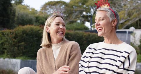 Two women laughing on backyard bench celebrating with festive tiaras and casual style