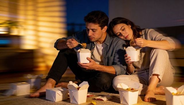 Couple enjoying cozy late-night takeout on rug sharing noodles with chopsticks, warm ambient lightin