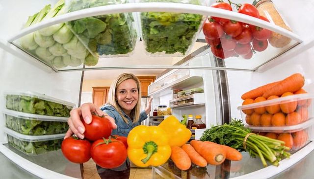Smiling woman reaching into fridge grabbing tomatoes among fresh vegetables and greens