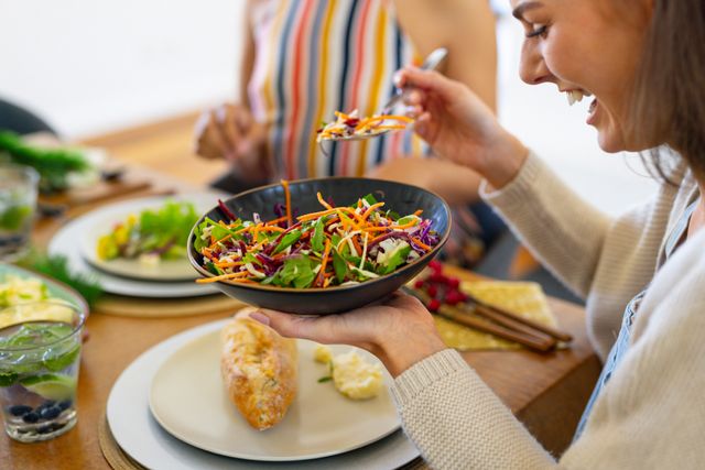 Woman serving herself a fresh salad while laughing at a dinner table with friends. Perfect for promoting healthy lifestyles, social gatherings, and the joy of eating together. Ideal for use in advertisements, blogs, and articles about nutrition, friendship, and home dining.