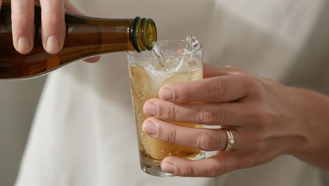 Pouring sparkling drink from amber bottle into tumbler by ringed hands close-up at home