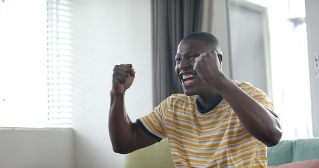 Excited african american man watching sport on tv at home celebrating victory, copy space. Lifestyle, sport, competition, entertainment and domestic life, unaltered.