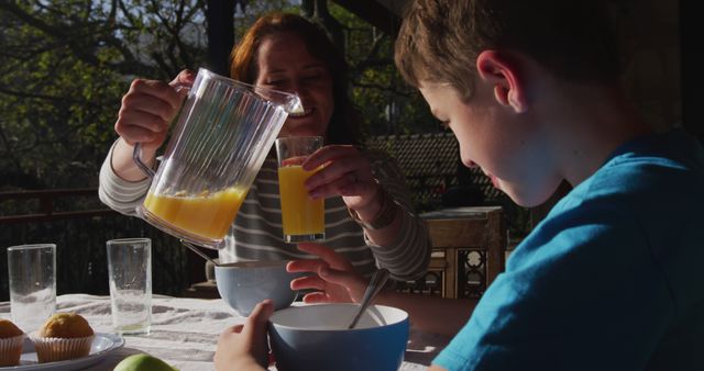 Mother pouring orange juice into her son's glass while sitting at a table outdoors enjoying breakfast. The morning sunlight creates a warm atmosphere, perfect for depicting family bonds and healthy lifestyles. Great for use in advertisements for family products, health foods, and outdoor dining experiences.