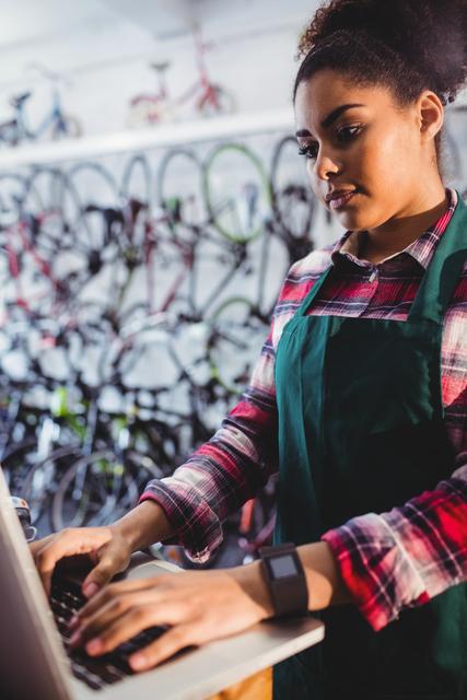 Young female mechanic in a bicycle workshop using a laptop for work. She is wearing an apron and standing in front of a wall filled with bicycles. This image can be used to depict modern technology in traditional professions, women in technical roles, or small business operations.