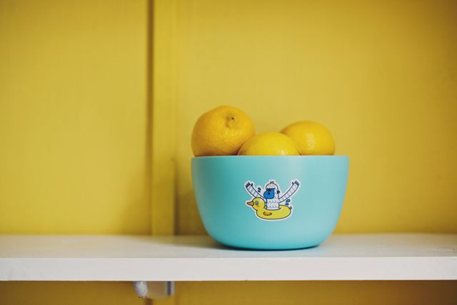 This colorful scene features a blue ceramic bowl filled with fresh lemons, set against a rich yellow background. The bowl is adorned with a playful cartoon character sticker, adding a whimsical touch. Perfect for illustrating concepts of freshness, simplicity, and vibrant kitchen decor. Ideal for magazines, websites, or blogs focusing on home living, interior design, food, and lifestyle.