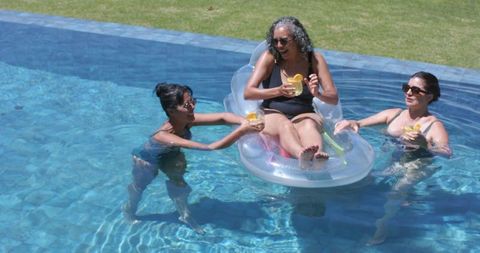 Women laughing and floating on inflatable pool lounge in sunlit backyard holding drinks