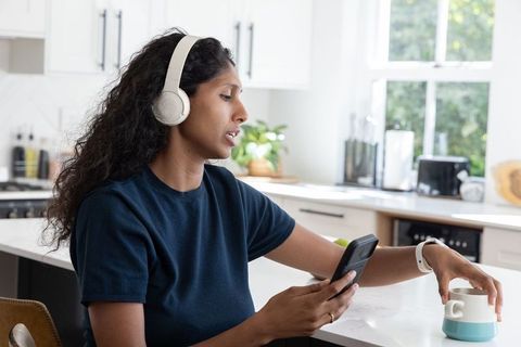 African american woman with headphones at kitchen counter checking phone reaching for mug