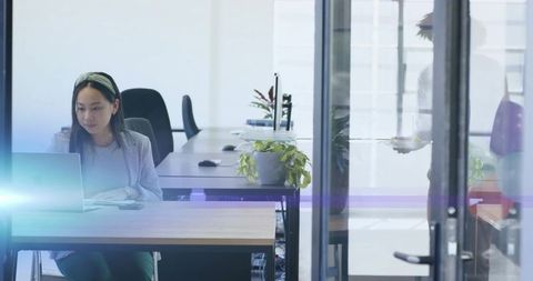 Asian woman typing on laptop at open-plan office desk with plants and glass partition