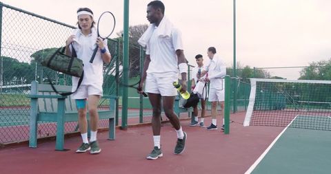 Young male tennis players walking off court carrying rackets, bags and water bottles