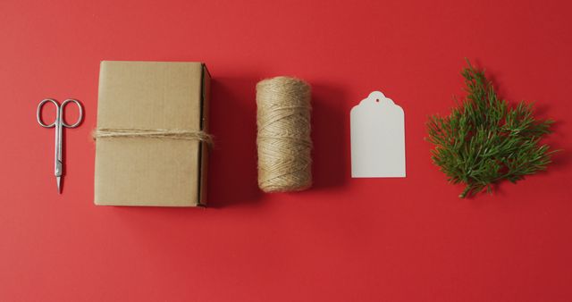Gift wrapping items neatly arranged on red background, featuring a cardboard box, twine, scissors, gift tag, and greenery. Ideal for holiday preparation, crafting guides, gift wrapping tutorials, and festive DIY projects.
